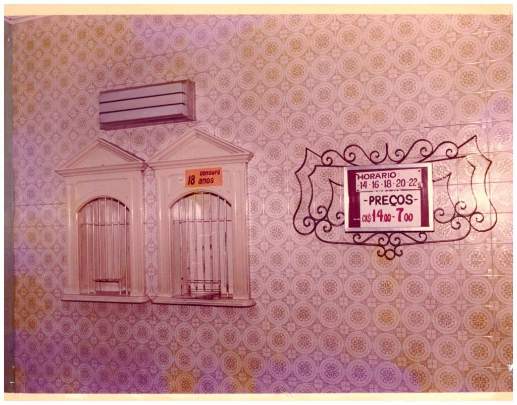 Fotografia retangular horizontal da bilheteria do Cine Capitólio. Década de 1980. Na parede estampada destacam-se duas bilheterias que tem ao lado informações sobre os horários e preços das sessões. [Fim da descrição]