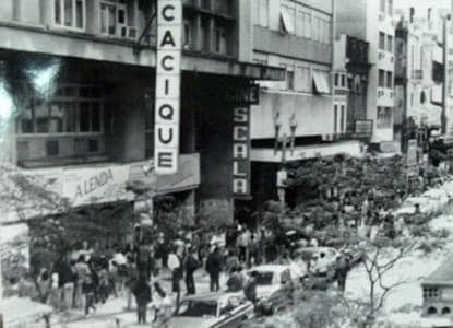 Fotografia retangular em preto e branco,   horizontal, dos cinemas Cacique e Scala. Década de 1980. Uma avenida com automóveis estacionados e muitos edifícios e  pessoas caminhando na calçada. Destacam-se letreiros dos cinemas Cacique e Scala. [ Fim da descrição]