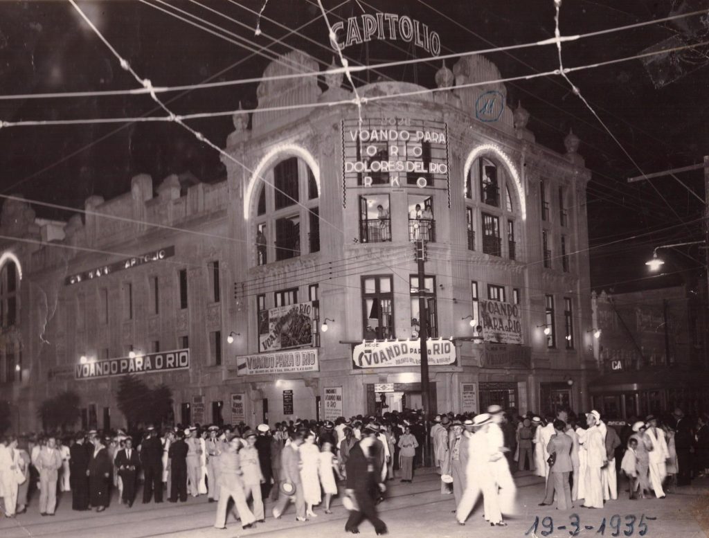 Fotografia em preto e branco retangular  horizontal do Cine Theatro Capitólio. Ano :1935. O prédio de esquina tem o letreiro iluminado  com a frase “ Voando para o rio” e  na calçada em frente  há pessoas  que formam uma fila e outras caminhando. [Fim da descrição]