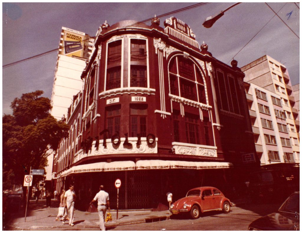 Fotografia retangular horizontal do Cine Capitólio. Década de 1980. O Prédio de esquina vermelho-escuro tem detalhes nas janelas em branco e o letreiro com a palavra Capitólio. Em frente ao prédio há um Fusca vermelho parado e algumas pessoas caminhando na calçada. [Fim da descrição]