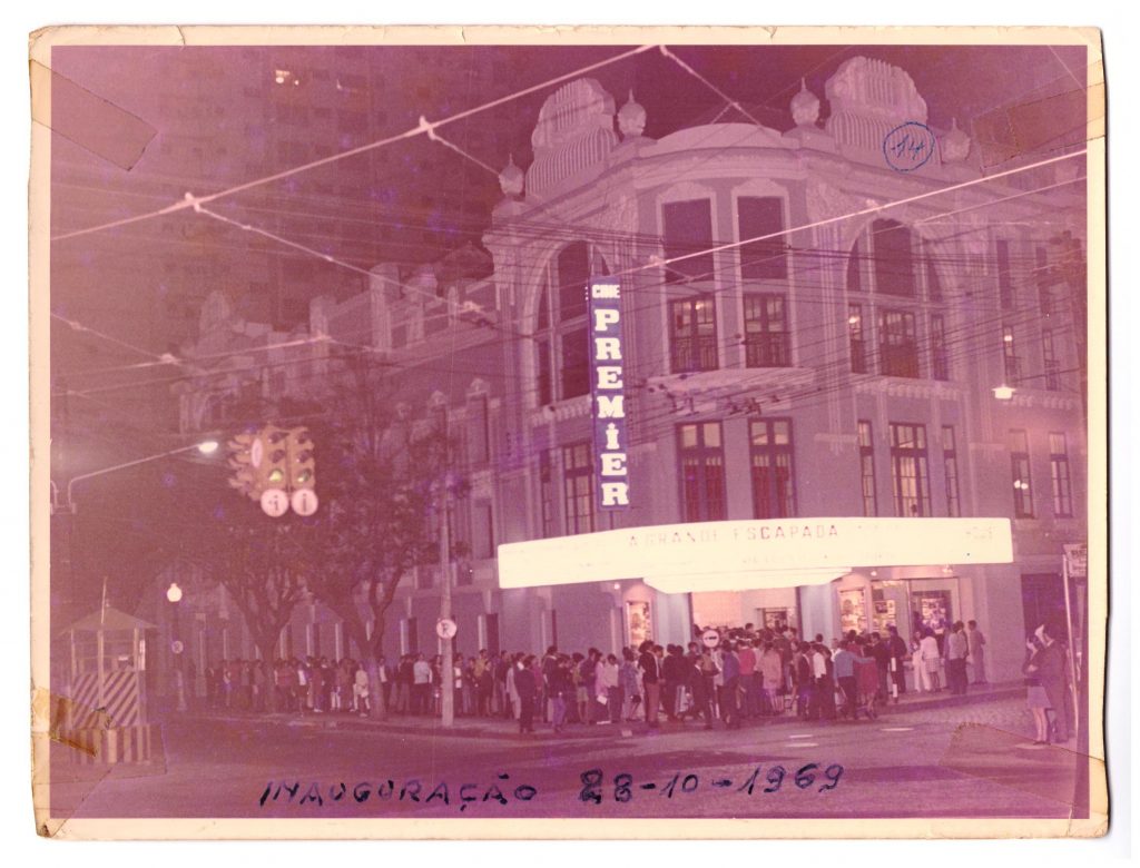 Fotografia em preto e branco retangular horizontal do Cine Premier. Ano : 1969. O prédio de esquina tem o letreiro iluminado com a palavra Premier. Na calçada em frente há pessoas em uma fila e outras caminhando. [ Fim da descrição]