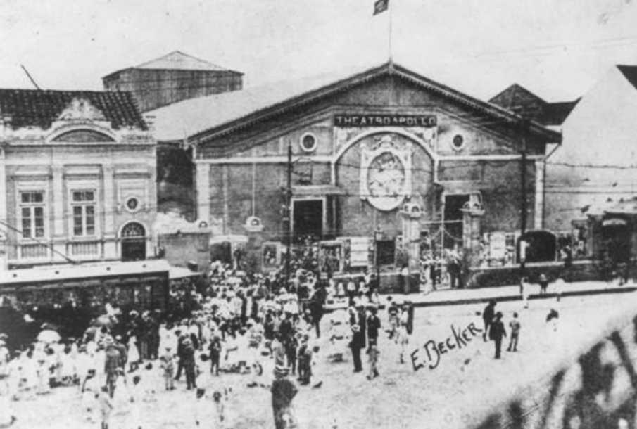 Fotografia em preto e branco retangular da fachada do Theatro Apollo, em 1914. O prédio do cinema eestá enquadrado ao centro com uma fila de pessoasno entorno para entrar no prédio. Fim da descrição.