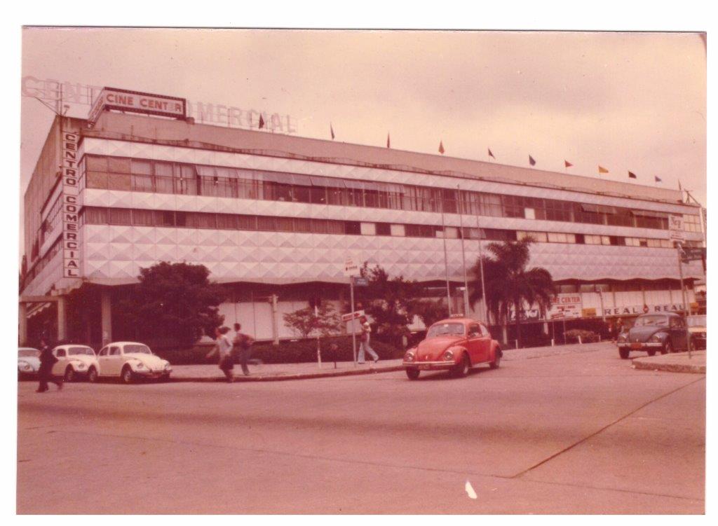 Fotografia retangular horizontal do Centro comercial João Pessoa. Década de 1970. Em primeiro plano, um cruzamento entre duas avenidas asfaltadas. Tem um táxi na cor laranja. Há vários automóveis estacionados na avenida e pessoas caminhando na calçada. Ao fundo, um prédio baixo ocupa toda extensão da imagem. Do lado esquerdo do prédio tem um letreiro vertical com a frase “Centro Comercial”. Em cima do prédio há um letreiro horizontal com a frase “Cine Center” e várias bandeirolas que tremulam ao vento.