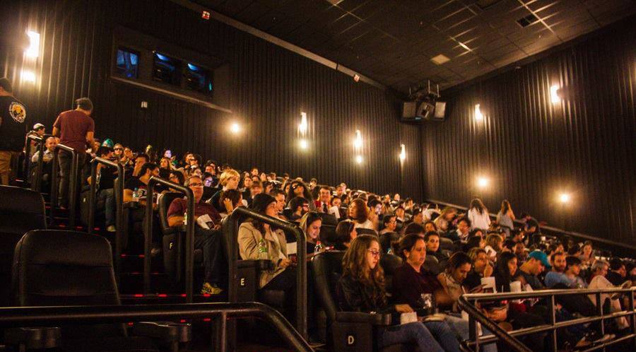 Fotografia retangular horizontal de uma sala de cinema. Ano: 2017. A sala tem paredes escuras e pouca iluminação. As poltronas enfileiradas formam uma inclinação, estão quase todas ocupadas. As pessoas que estão sentadas comem, bebem refrigerante, olham seus celulares, olham ao longe.  Fim da descrição