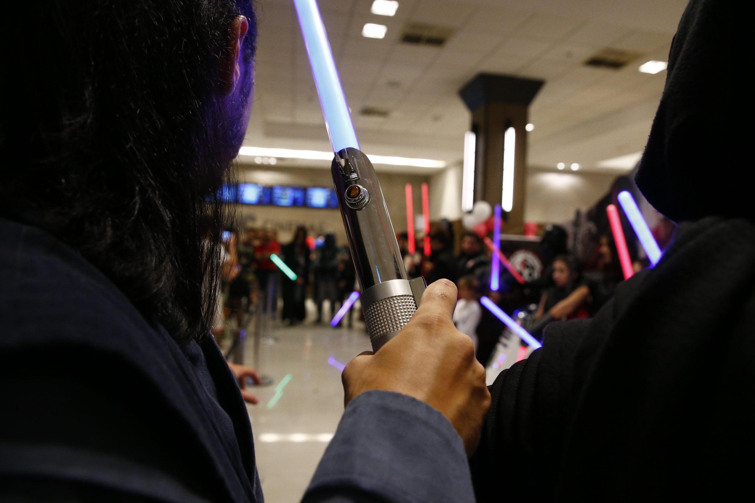 Fotografia retangular horizontal   da pré-estreia do filme Os últimos Jedi. Ano: 2017. Em primeiro plano, duas pessoas estão de costas, dos ombros para cima. A pessoa da esquerda tem cabelos longos, lisos e veste roupa cinza. No centro da imagem, uma mão segura um sabre de luz. A pessoa do lado direito usa capa com capuz. Ao fundo, há uma fila com várias pessoas segurando sabre de luz, que são coloridas.  Fim da descrição
