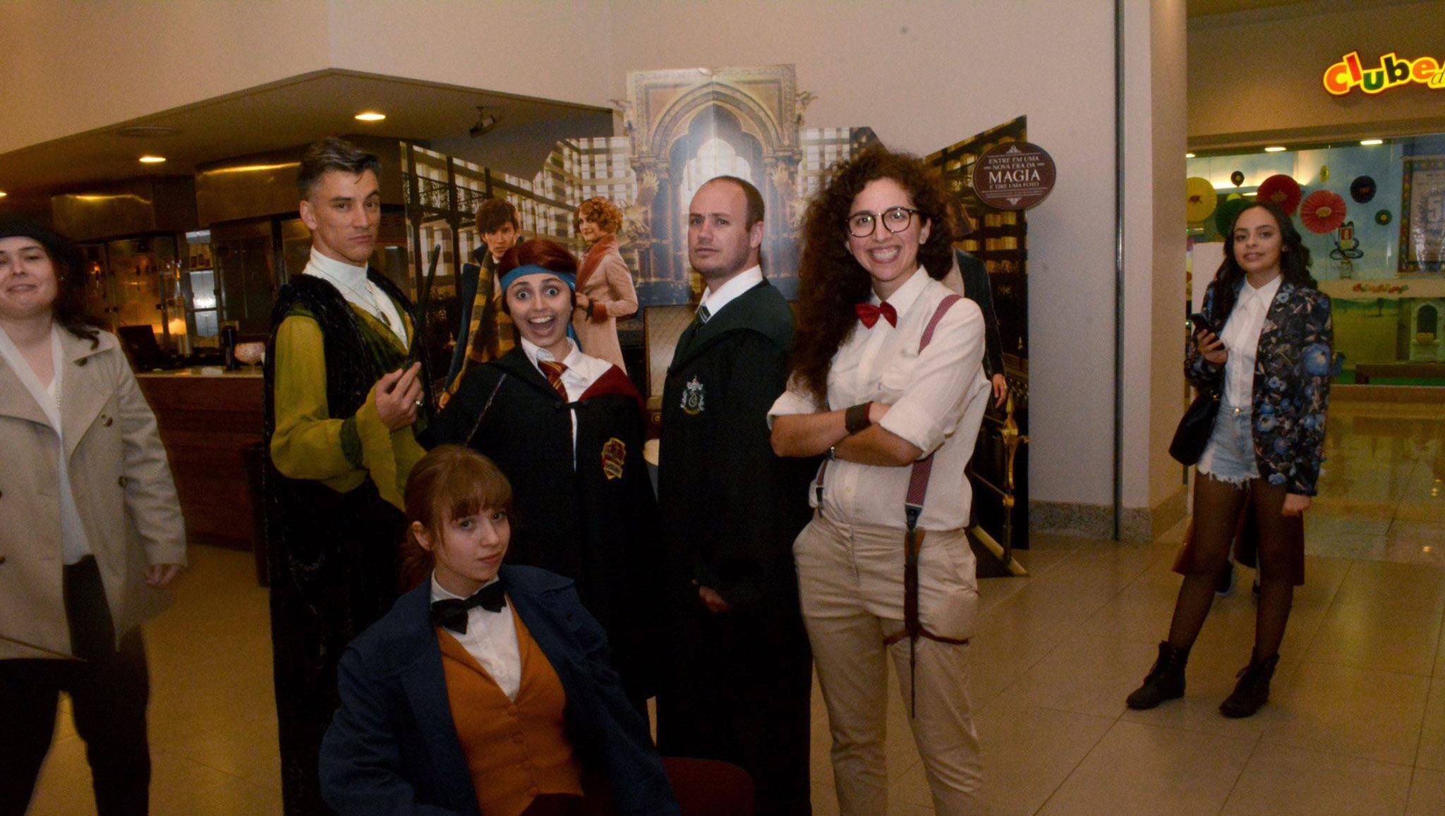 Fotografia retangular horizontal do Grupo Alohomora. Ano: 2017. Cinco pessoas estão em frente a uma sala de cinema usando vestimentas alusivas aos personagens do filme Harry Potter. Um homem branco, alto, tem cabelos pretos, está sério de perfil para a direita, este roupa preta com as mangas cor de mostarda e gola branca. Ao lado do homem, uma mulher branca, baixa de cabelos curtos tem uma fita azul na cabeça, está de frente, sorridente com a boca aberta, veste roupa preta caída no ombro que deixa à mostra a blusa branca. Ao lado da mulher, um homem branco de estatura mediana, tem cabelos curtos e pouca barba, sério, está de perfil para a esquerda, veste roupa preta com gola branca aparente. Ao lado do homem, uma mulher branca, alta, tem cabelos longos e crespos, usa óculos, está de frente, sorridente com os braços cruzados, veste calça bege, camisa branca com mangas até o cotovelo, suspensório marrom e gravata borboleta vermelha. Uma mulher branca está abaixada em frente ao grupo. Aparece da cintura para cima, com expressão séria, tem cabelos ruivos com franja, que estão presos. Veste casaco preto sobre blusa branca, colete cor de mostarda e gravata borboleta preta. Fim da descrição