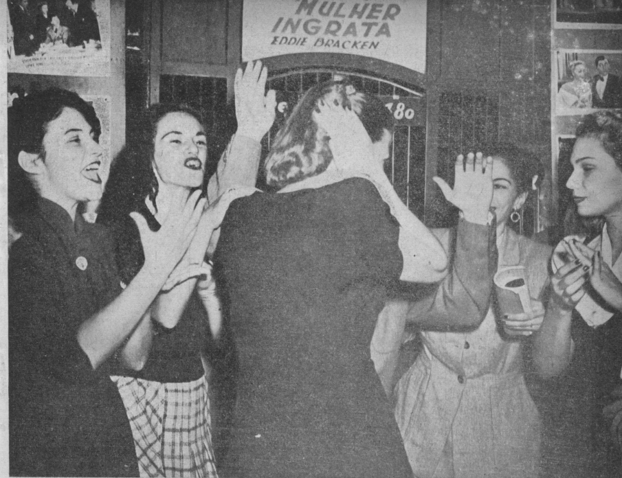 Fotografia em preto e branco tirada em frente à bilheteria do cinema. Acima, ao centro da imagem, os textos “mulher ingrata” e “Eddie Bracken”.  Nas extremidades laterais, fotografias de cenas de filmes. Ao centro da imagem, em primeiro plano, mulher de costas tapando os ouvidos e, em sua frente, quatro mulheres gritando e batendo palmas. [Fim da descrição]