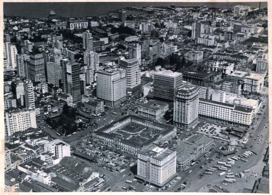 Fotografia em preto e branco, plano aéreo do centro da Cidade de Porto Alegre, com destaque ao centro da imagem para os prédios do Mercado Público, Prefeitura de Porto Alegre, Palácio do Comércio, e Edifício União, entre outros. [Fim da descrição]
