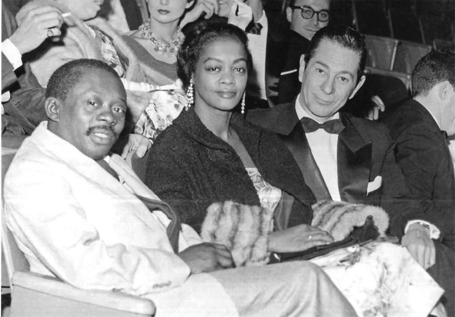 Imagem em preto e branco mostra sentados em poltronas de cinema, pela ordem, da esquerda à direita: em primeiro plano, o ator Grande Otelo, vestindo casaco em tonalidade clara e gravata escura; ao seu lado, a atriz Ruth de Souza, com casaco de tom escuro e peles nas mangas, além de brincos de pérolas; e, ao seu lado, o ator Oscarito, vestindo smoking em tom escuro, camisa clara e gravata borboleta em tom escuro. Ao fundo, sentados na fila anterior, quatro espectadores. [Fim da descrição]