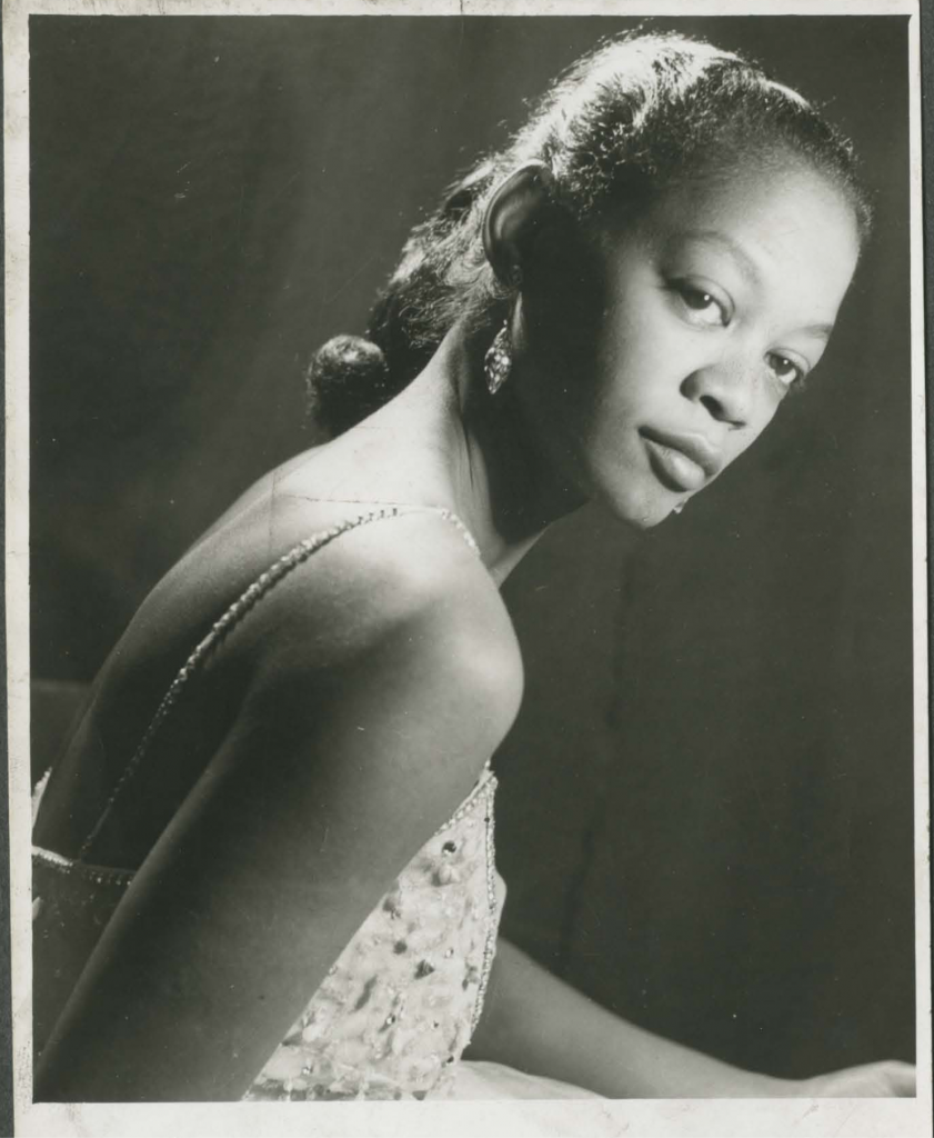 Fotografia em preto e branco, em modo retrato, da atriz Ruth de Souza. Nela, a atriz aparece em perfil, com as costas e antebraço em primeiro plano, trajando vestido de renda em tons claros, alças com pérolas e brinco brilhante, com os cabelos presos.[Fim da descrição]