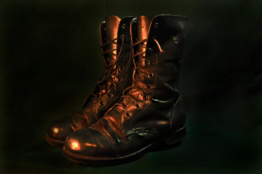 Fotografia retangular horizontal. Ao centro da imagem há um par de botas de couro de cor preta. A bota tem cano médio e cadarços que acompanham quase toda a extensão do cano da bota. O fundo da imagem é em outro tom de preto, o que confere destaque a bota em primeiro plano. [Fim da descrição]