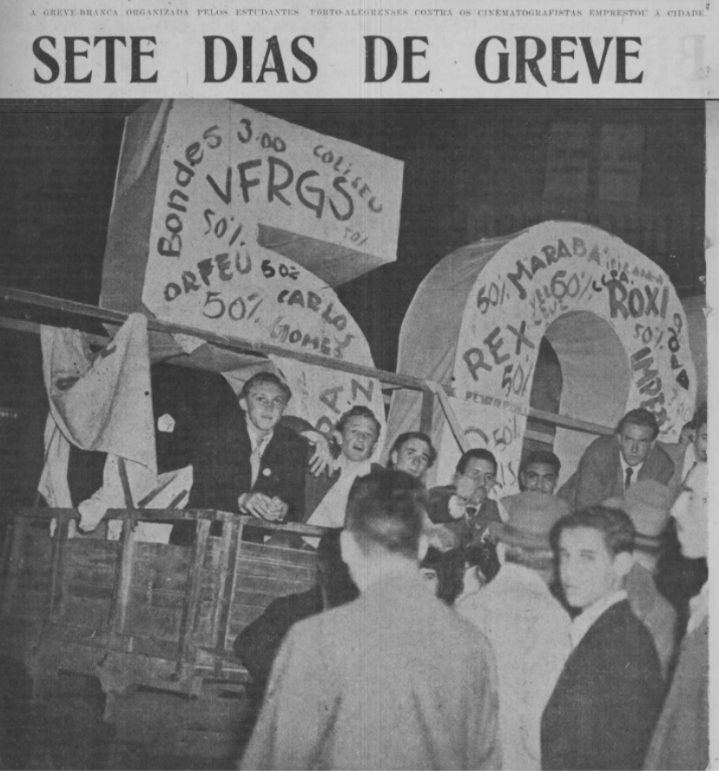 Fotografia em preto e branco, no centro de Porto Alegre,  em 1947 , seis jovens estão  em cima de um caminhão, todos bem agasalhados, em cima do caminhão também está uma  alegoria feita com tecido branco, com os números 5 e 0, que representa os  50%%%%, no número cinco está inscrito  (UFRGS)  Universidade Federal do Rio Grande do Sul, e no número 0 está inscrito o nome de alguns cinema como: rex, roxi e marabá. Na calçada em frente ao caminhão está uma multidão de  jovens também bem agasalhados, alguns com  chapéus na cabeça. [Fim da descrição]