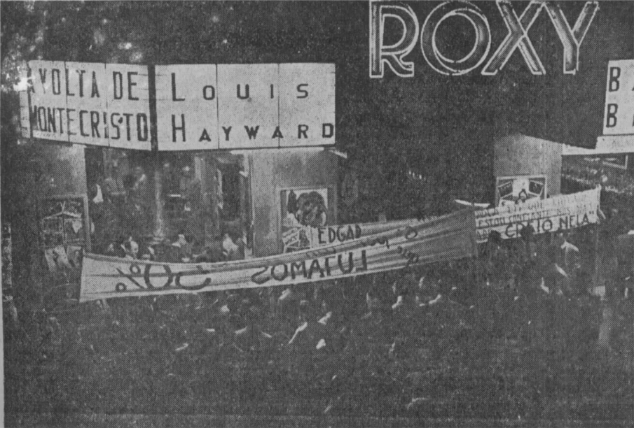 Fotografia em preto e branco no centro de Porto Alegre, em 1947 , uma  multidão em frente ao cinema, que tem um letreiro iluminado com o nome filme “A volta de monte cristo”. [Fim da descrição]