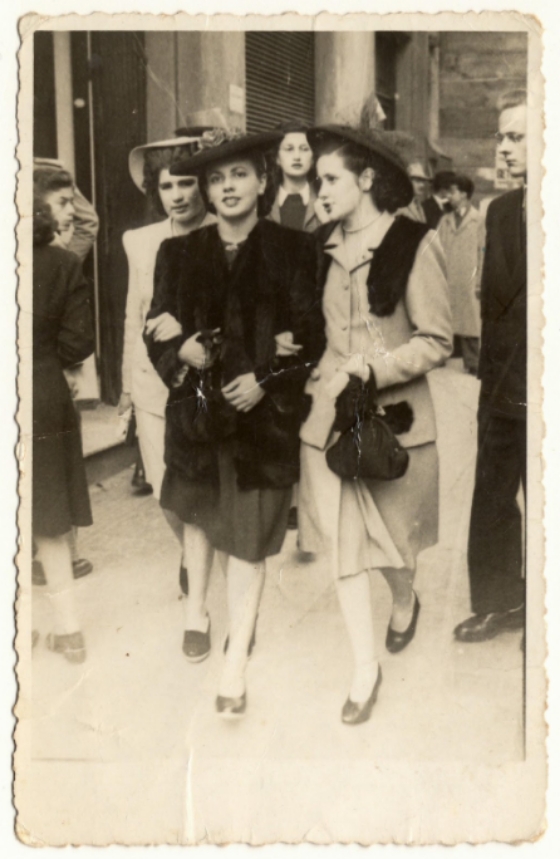 Fotografia no formato retrato em tons de sépia, de 1940. No centro da imagem duas mulheres passeiam pela Cinelândia de Porto Alegre. A mulher à esquerda veste traje escuro. A mulher à direita veste trajes claros e seu rosto está virado para a mulher à esquerda. Ambas usam chapéus e carregam bolsas de mão. Caminham com os braços dados. O fundo da imagem é a Cinelândia porto-alegrense, composta por vários transeuntes. [Fim da descrição].