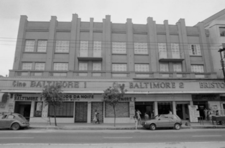 Fotografia em preto e branco retangular,  horizontal do Cinema Baltimore 1 e 2. Década de 1980. Em primeiro plano  uma avenida com dois carros estacionados e  poucas pessoas caminhando na calçada. Ao fundo o prédio do Cinema Baltimore 1 e 2. [Fim da descrição]