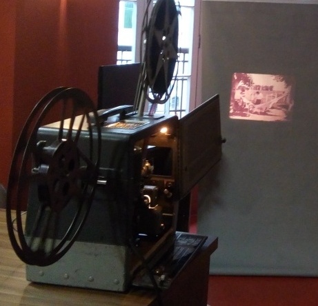 Fotografia quadrada de um projetor de filmes. Década de 1980. O equipamento está ligado e projeta filme na parede . [Fim da descrição]