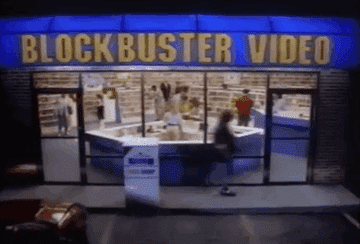 Imagem  retangular, horizontal  da fachada  da vídeo locadora Blockbuster Vídeo. Letreiro com fundo azul e letras amarelas. As  pessoas estão em movimento, saem e entram na loja.  No interior as prateleiras estão cheias de filmes e de pessoas caminhando. [Fim da descrição]