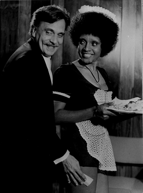 Imagem em preto e branco  vertical de uma cena do filme “Como é boa nossa empregada” com Aizita Nascimento e Jorge Dória. Na cena uma jovem mulher negra está em pé  de perfil direito. Ela usa vestido escuro de mangas curtas e avental branco. Segura uma bandeja com as duas mãos na altura do abdômen. Atrás dela está um homem branco, de terno escuro que encosta a mão direita  na perna dela. Os dois sorriem. [Fim da descrição]