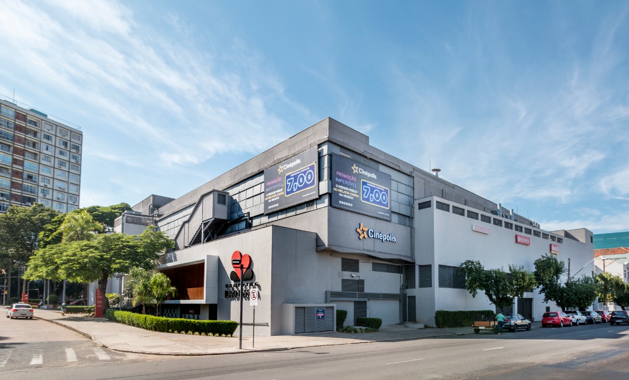 Fotografia retangular horizontal do Centro comercial João Pessoa. Década de 2020 Em primeiro plano, um cruzamento entre duas avenidas asfaltadas têm automóveis estacionados em ambas. No centro da imagem, o prédio baixo ocupa toda a quadra, que tem canteiros e árvores na calçada. O Prédio tem muitos logotipos coloridos nas paredes externas, inclusive da empresa de cinemas do local. Fim da descrição